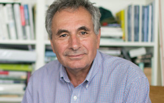 College Awards Honorary Doctorate To Ron Phillips A Pioneer Of Community Economic Development In Maine 5 A man with gray hair and a plaid shirt is sitting in front of a bookshelf filled with books and binders. He is looking directly at the camera, evoking Ron Phillips' dedication to Community Economic Development. Saint Joseph's College of Maine