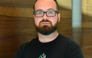 Redefining The Fitness Business 10 A man with a beard and glasses is wearing a black t-shirt with a logo that reads "ASF - Pod Health." He is standing against a wooden background, embodying the spirit of redefining fitness within the fitness business. Saint Joseph's College of Maine