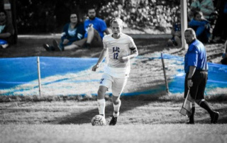 Chasing The Competitive Edge On The New Turf Field 7 A soccer player in a white jersey with the number 12 dribbles the ball on a pristine turf field. Spectators and an official are visible in the background, adding to the competitive edge of the game. Saint Joseph's College of Maine