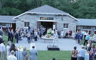 Shaping Maine 9 A gathering of people is taking place outside a stone building with large open doors. Attendees, key players in the Shaping Maine initiative, are standing and mingling on a paved courtyard, with some seated at tables on the lawn. Saint Joseph's College of Maine