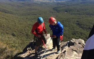 Classroom: Beyond Four Walls 6 Two rock climbers wearing helmets and harnesses stand near the edge of a cliff, overlooking a vast forested landscape beyond the confines of four walls under a clear sky. Saint Joseph's College of Maine