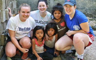 Serving The Common Good At Saint Joseph’s College 1 Three women and three children, embodying the spirit of serving the Common Good, pose for a photo in front of a partially constructed or damaged building. The women are squatting, and the children are sitting on the ground. Saint Joseph's College of Maine