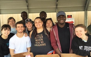 Spring Break Workfest: Student Service Trips During Lent 9 A group of smiling people stand under a shelter, some wearing casual clothing and one with a shirt that reads "Life is better when you're laughing." They appear to be students on a Spring Break service trip, volunteering at an event. Saint Joseph's College of Maine