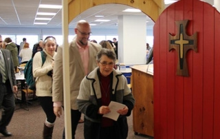 Memorializing Mercy 6 People are walking through an archway with a wooden door that has a cross on it, evoking a sense of mercy. In the foreground, a woman is holding papers, memorializing those who came before her, while others follow behind her. Saint Joseph's College of Maine