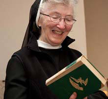 Sister Mary De La Salle O’donnell Retires 3 Sister Mary, an elderly woman in a nun's habit, smiles while holding and looking at a book with a green cover, enjoying her quiet moment of reflection in retirement. Saint Joseph's College of Maine