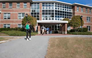 Students walking towards an academic building with a sign reading "Freeden Hall" on a sunny day, embracing the camaraderie that comes with communal living. Saint Joseph's College of Maine