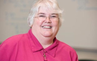Dr. Susan Miovech ’82 2 A person with short white hair wearing glasses and a pink top smiles at the camera, reminiscent of Dr. Susan Miovech, a revered 1982 alumni. Saint Joseph's College of Maine