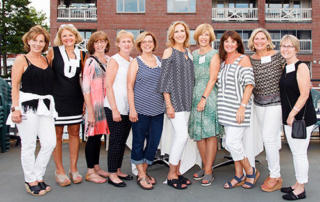 Getting On Board 3 A group of ten women standing in a row outdoors, wearing casual summer outfits, getting on board with the spirit of the season. A brick building is visible in the background. Saint Joseph's College of Maine