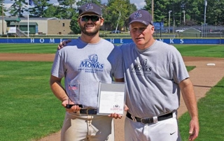 Hitting The Books 5 Two men in matching shirts and baseball caps stand on a baseball field holding an award and a certificate. The man on the left is smiling with his trophy, while the other has his arm around him. It looks like their hard work both on the field and hitting the books has truly paid off. Saint Joseph's College of Maine