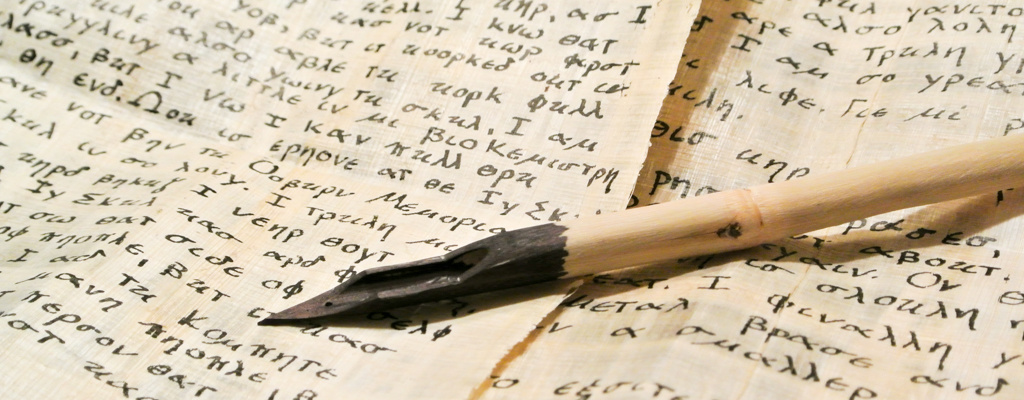 A wooden quill with black ink rests on parchment papers filled with handwritten text in Greek, likely the meticulous work of the Theology faculty. Saint Joseph's College of Maine