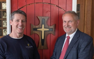 Rooted In Mercy 1 Two men pose together in front of a wooden wall featuring a cross, symbolizing their rooted faith. One man is wearing a navy sweatshirt, and the other is in a suit and red tie. Saint Joseph's College of Maine