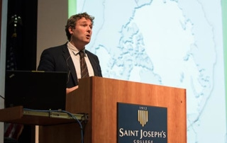 Ties To The Arctic 6 A man speaks at a lectern with a "Saint Joseph's College" sign; a large map of the Arctic is displayed on a projector screen behind him. Saint Joseph's College of Maine