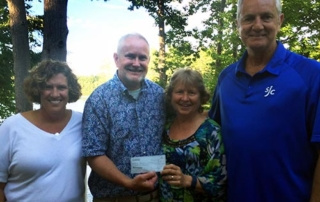 The John Waldron Scholarship 2 Four people are standing outdoors among trees with a lake in the background. The two in the middle, recipients of the John Waldron Scholarship, are proudly holding a check. Saint Joseph's College of Maine