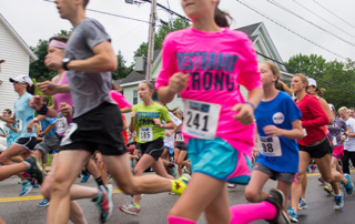 Going Strong In Westbrook 5 A group of runners, including a girl in a bright pink "Going Strong" shirt, participate in a race on a street lined with houses. Saint Joseph's College of Maine