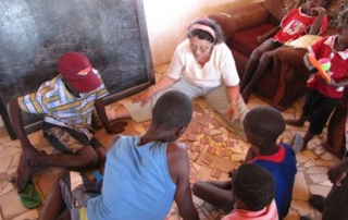 It’s A Long Way To Zambia, But Not When You Travel With Heart 3 An adult sits on the floor with a group of children, playing a card game in a room with a tiled floor and a blackboard on the wall, their laughter and joy reflecting the heart of Zambia's vibrant spirit. Saint Joseph's College of Maine